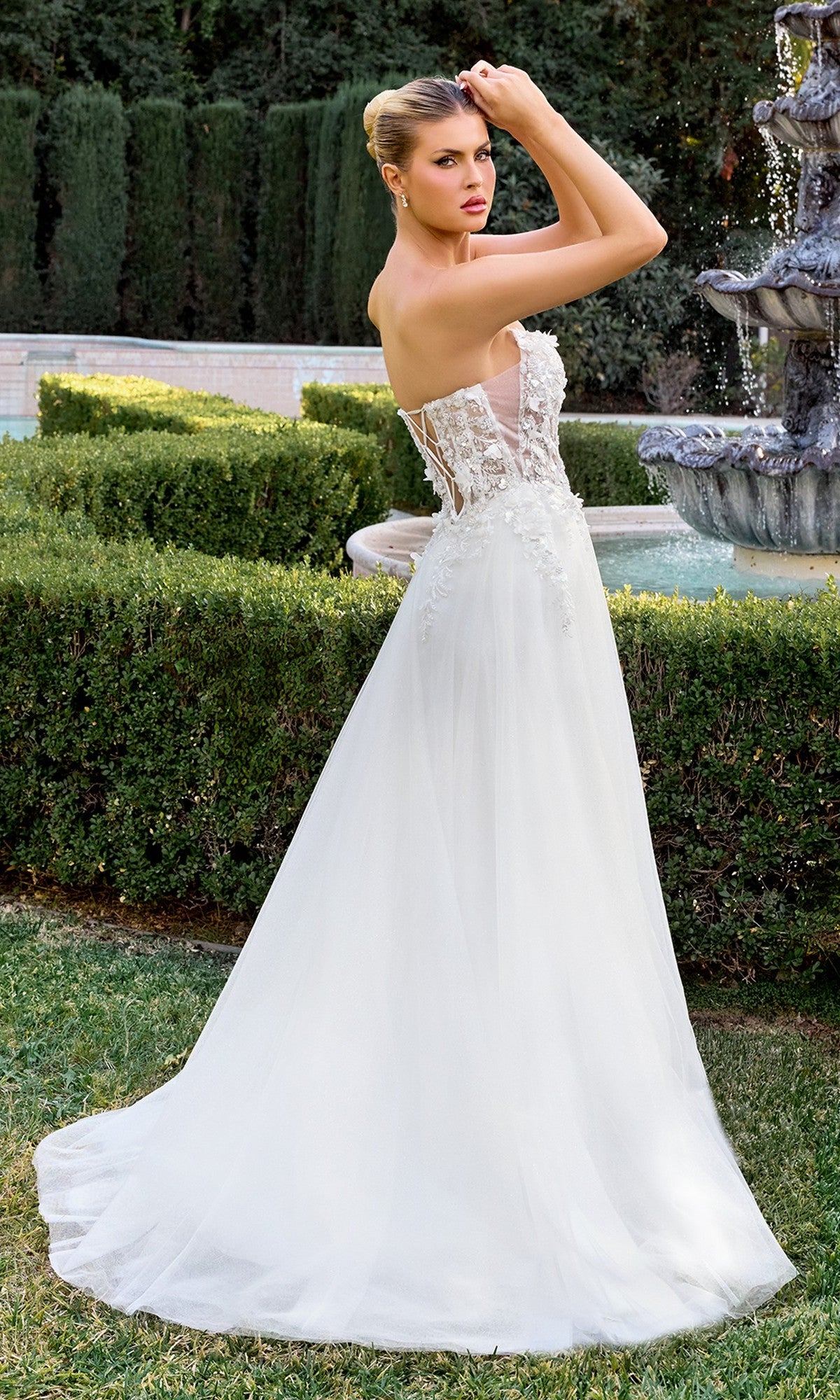 Wearing the Ladivine C174W white formal dress with a strapless sheer lace bodice and 3D floral appliqué, a woman stands outdoors by a fountain and manicured hedges, gazing over her shoulder.